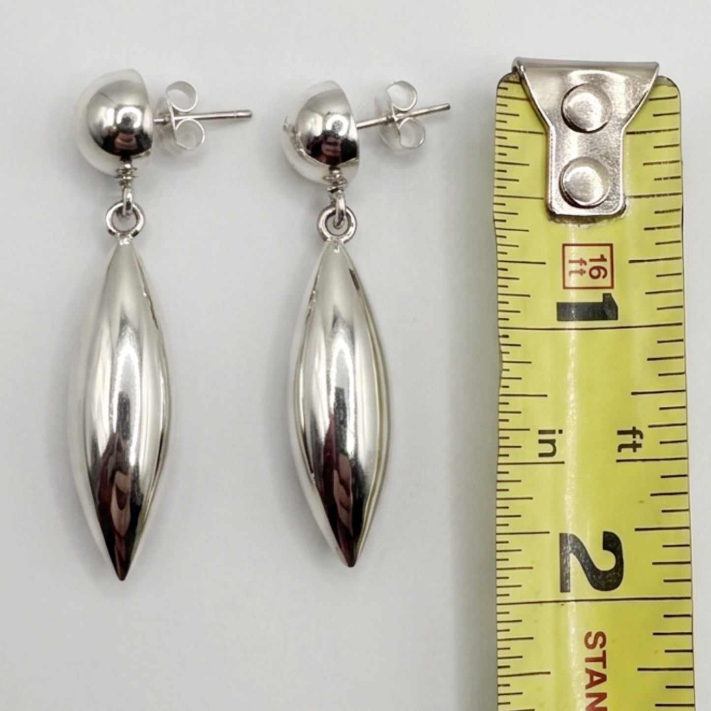 A pair of polished silver drop earrings with teardrop-shaped pendants and ball studs, displayed next to a yellow measuring tape to show their size against a light gray background.
