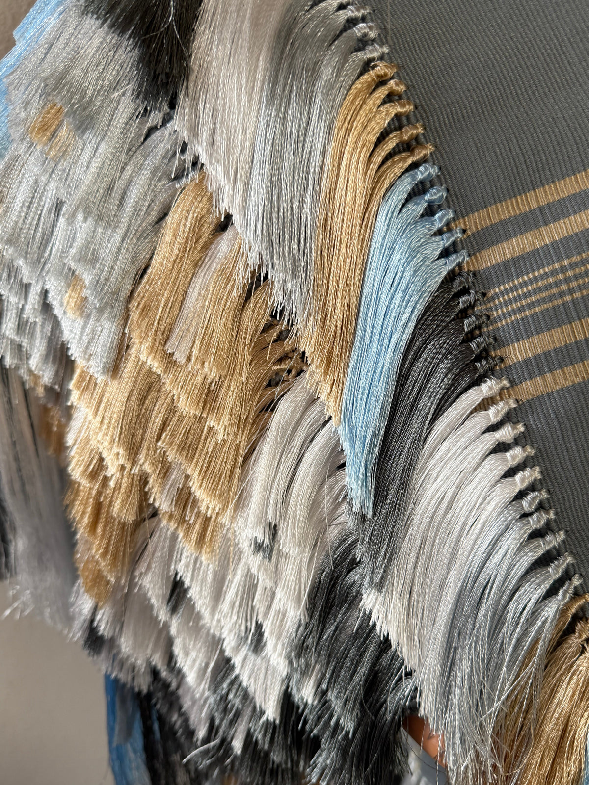 A close-up of a woven textile with layered fringe tassels in gray, white, beige, black, and light blue, with a gray base showing subtle gold stripes.