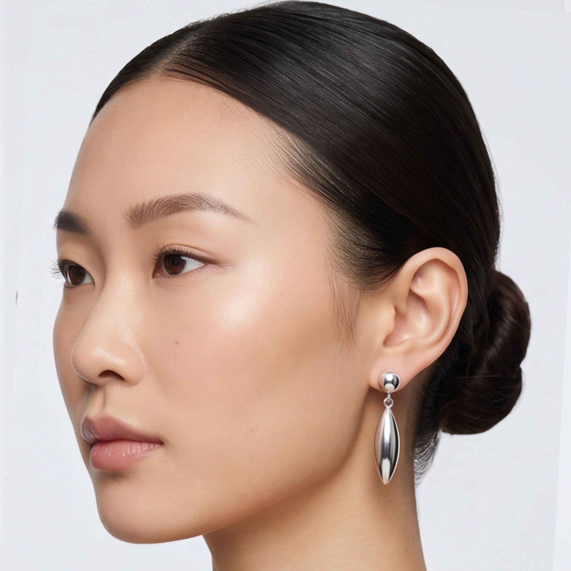 A close-up side profile of a woman with dark hair in a low bun, wearing a single silver teardrop-shaped drop earring against a light gray background.
