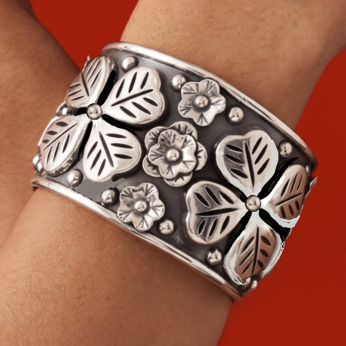 A wide silver cuff bracelet with raised floral and leaf motifs, worn on a person's wrist against a solid red background.