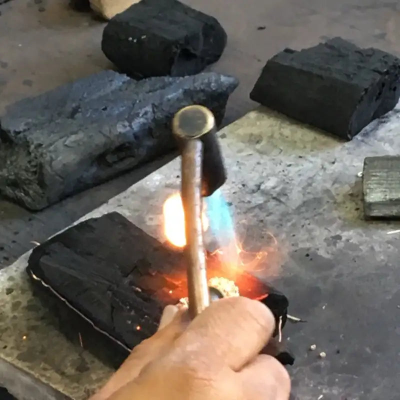 A brass hammer with a rounded head is being used to heat a piece of metal over glowing charcoal.