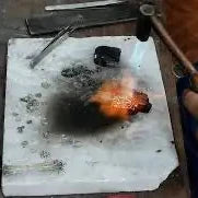 A person is using a torch to melt and shape an orange-colored metal piece on a white workbench.