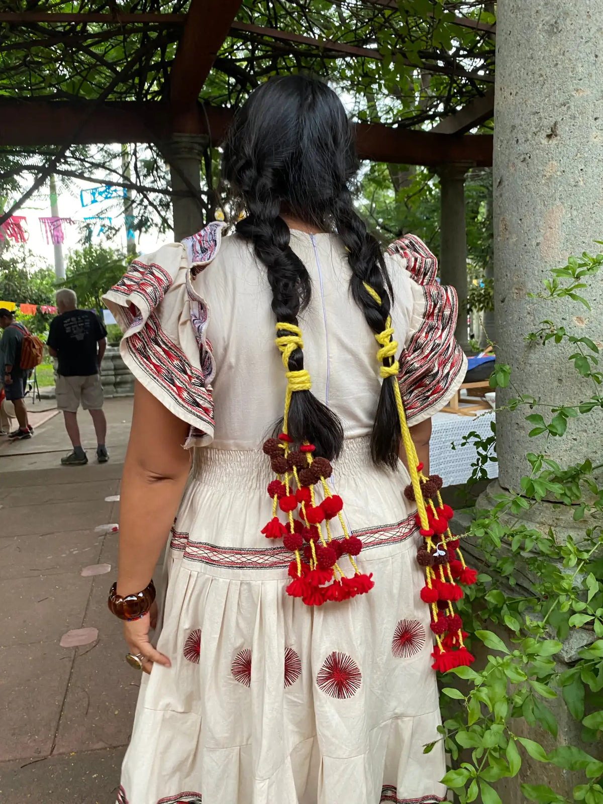 A woman wearing a cream-colored dress with red embroidered patterns and ruffled sleeves, her long black braids adorned with yellow ties and hanging clusters of red pom-poms.